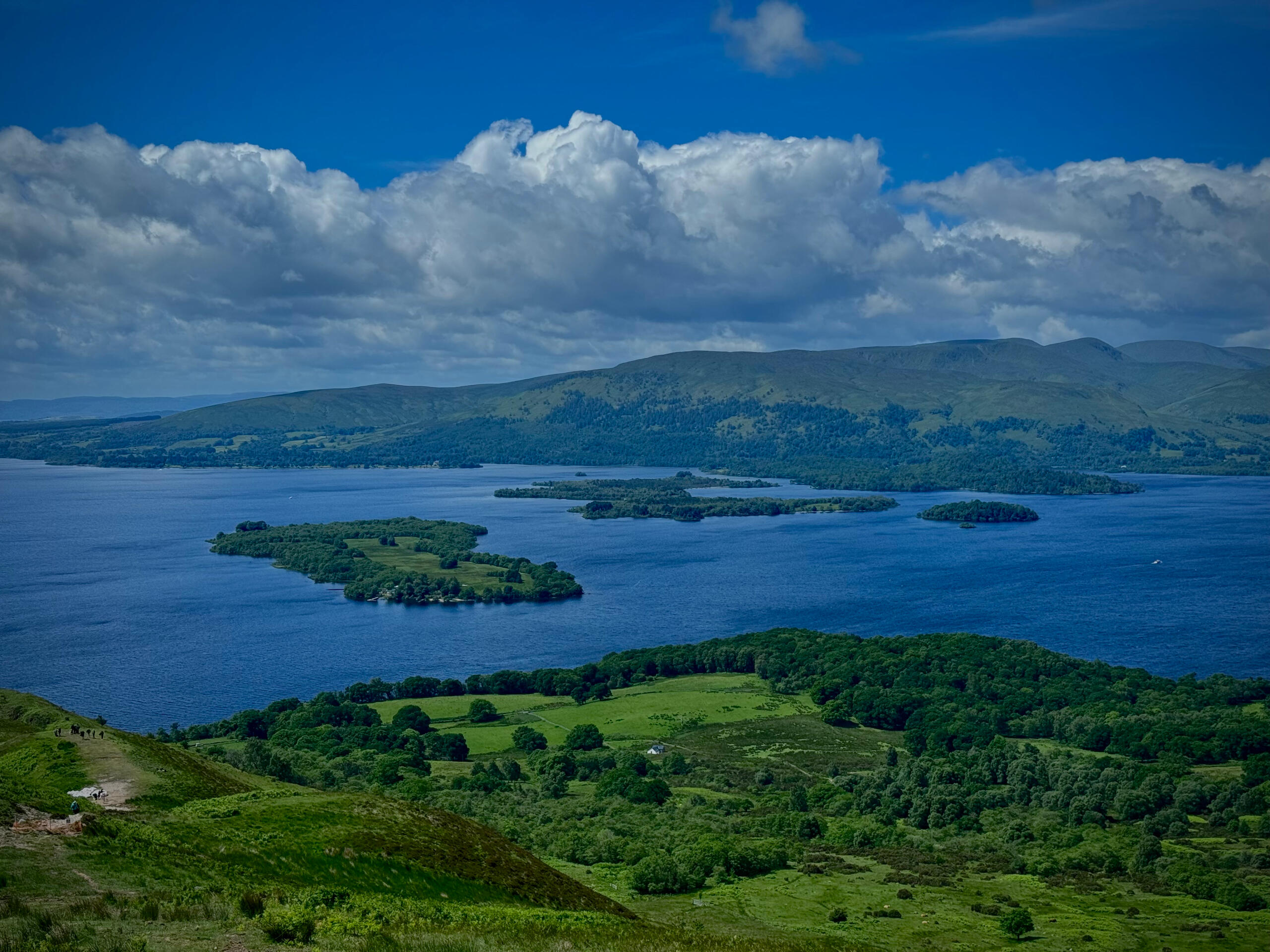 A stunning view of a Loch Lomond during summer with lush greenery, blend sky and fluffy clouds. Captured with iPhone 15 Pro.